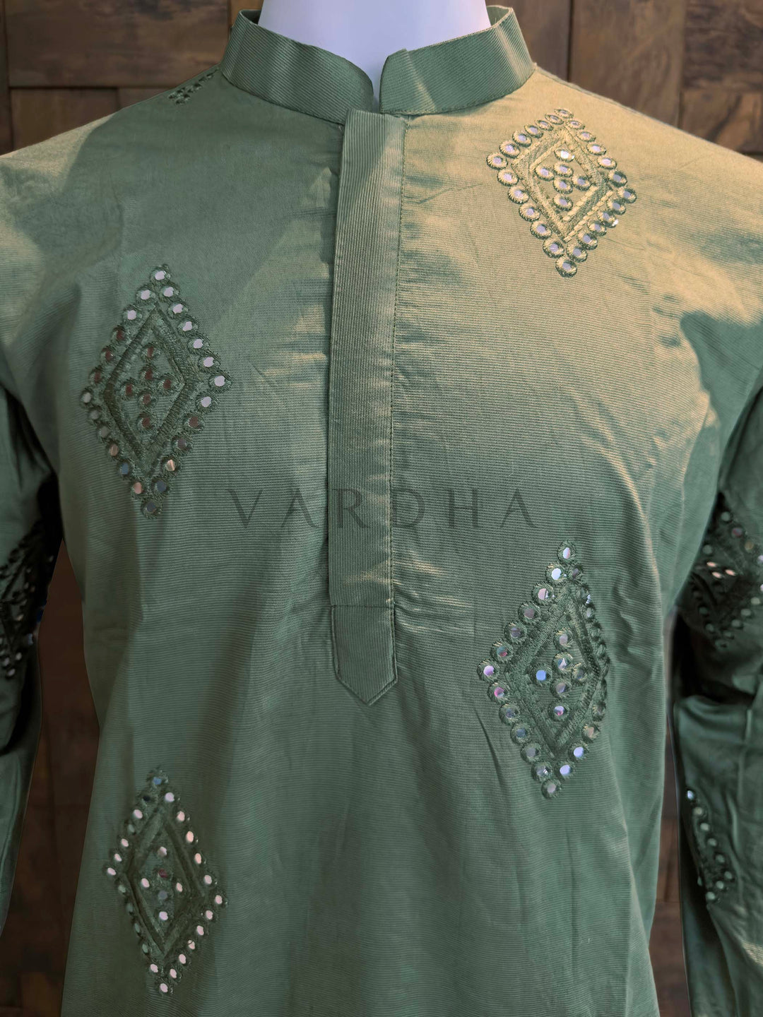 Green embroidered shirt on a mannequin with 'Vardha' branding. Close-up of sage green kurta neckline showing neat band collar, clean placket construction, and reflective mirror embroidery in diamond motifs.