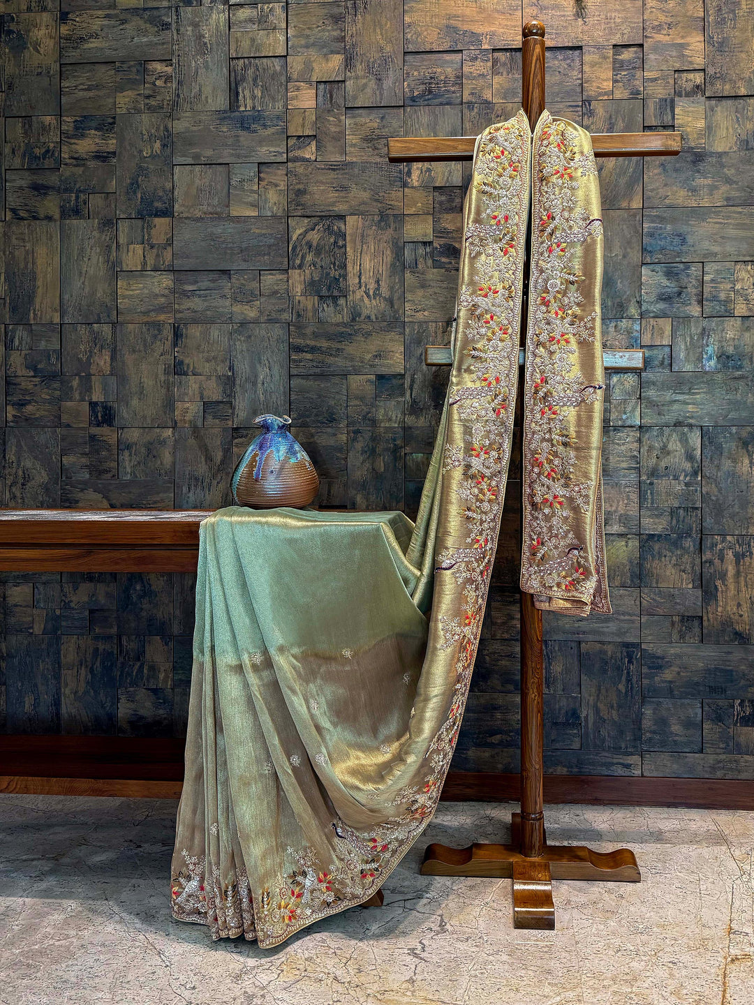 Dual-tone gradient body view of saree flowing from light sage to antique gold with light thread detailing. Decorative robe with floral and paisley patterns on a wooden surface