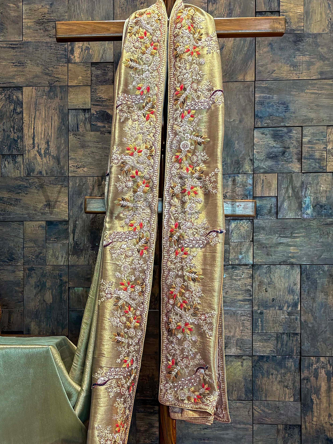 Close-up of tissue silk saree border featuring detailed peacock motifs and floral vines embroidered with zardozi, cutdana, and bead work. Decorative gold fabric with intricate patterns on a wooden stand against a textured wall.