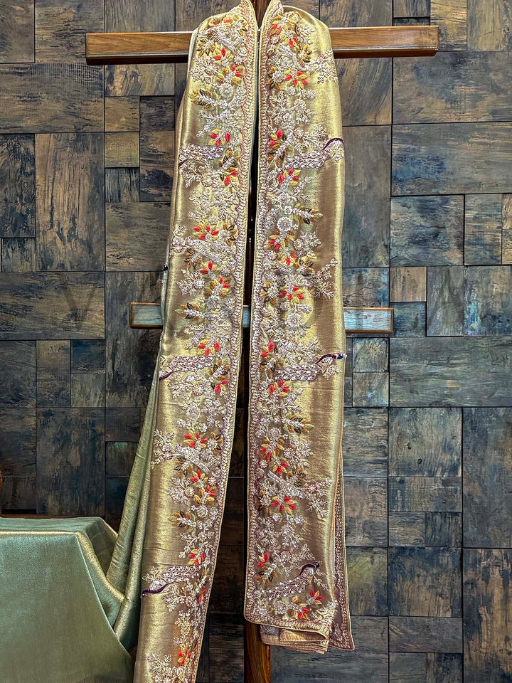 Close-up of tissue silk saree border featuring detailed peacock motifs and floral vines embroidered with zardozi, cutdana, and bead work. Decorative gold fabric with intricate patterns on a wooden stand against a textured wall.