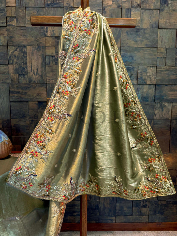Decorative green fabric with intricate embroidery on a wooden stand against a textured wall. Lower pleats displaying subtle tissue sheen and delicate scattered floral thread motifs near the hemline.