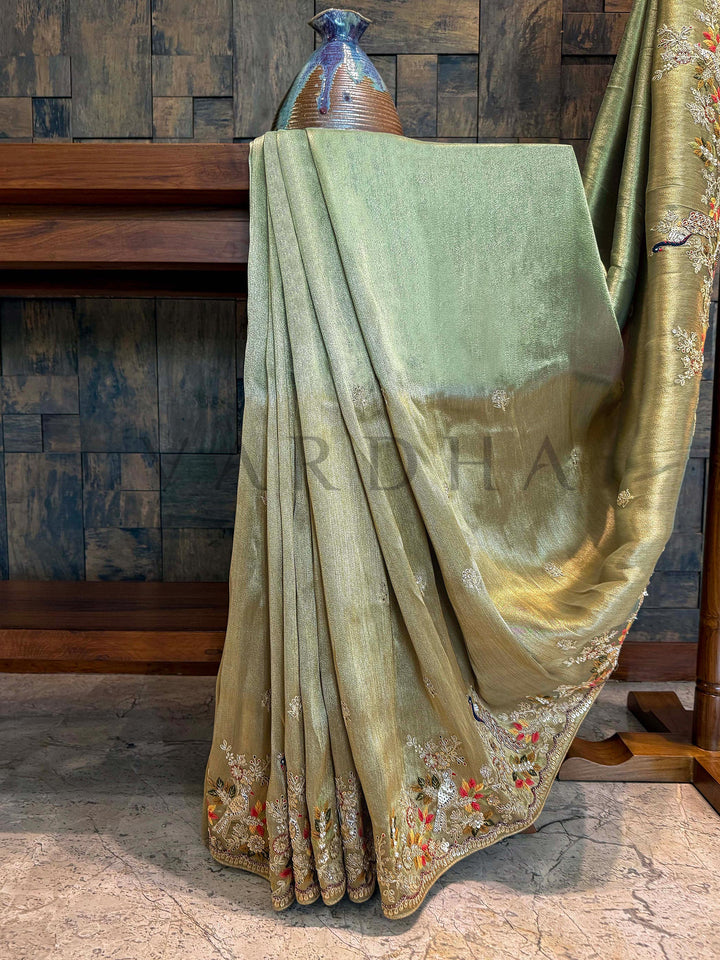 Decorative robe with floral patterns on a wooden cross against a textured wall. Rich zari tissue texture of sage green saree paired with vibrant embroidery showcasing regal craftsmanship.