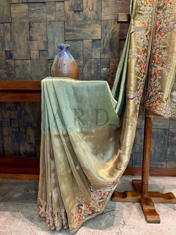 Pallu showcasing full-length hand embroidery with multicolored vines and peacocks on a dual-tone green-gold tissue base. Decorative fabric draped over a chair with a textured wall background