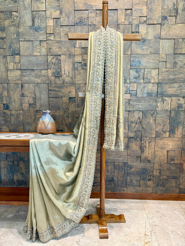 Saree draped over a wooden stand against a stone wall background Model showcasing sage green saree with elegant drape and shimmering zari tissue fabric.