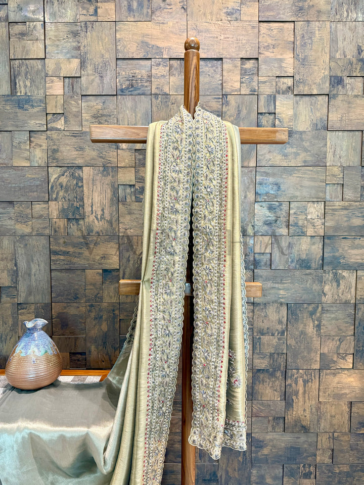 Long, patterned Saree draped over a wooden hanger against a wooden paneled wall. Sage green tissue silk saree featuring intricate floral embroidery with sequin and beadwork, draped gracefully.