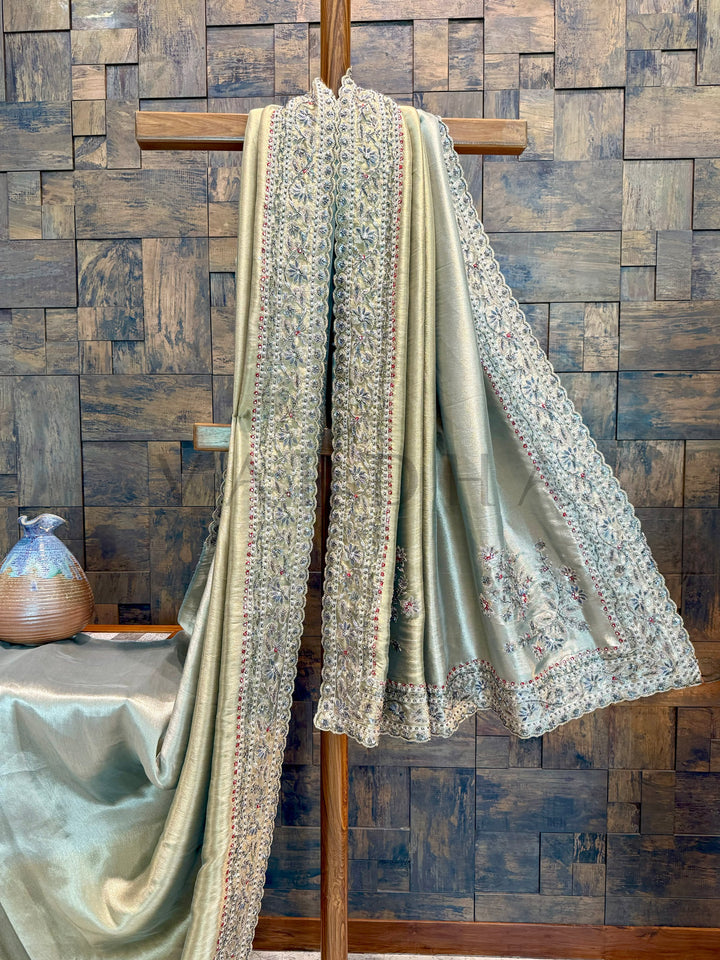 Light-colored fabric with intricate patterns draped over a wooden rod against a wooden paneled wall. Matching blouse piece with border-ready detailing perfect for styling and customization.
