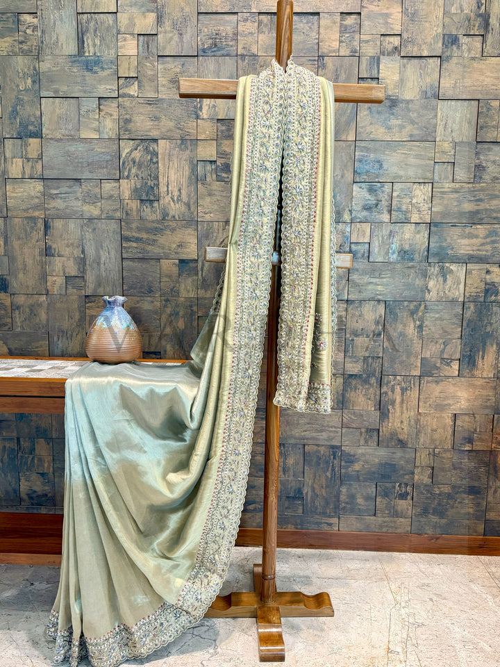 Light green saree with silver border draped over a wooden stand against a wooden paneled wall.