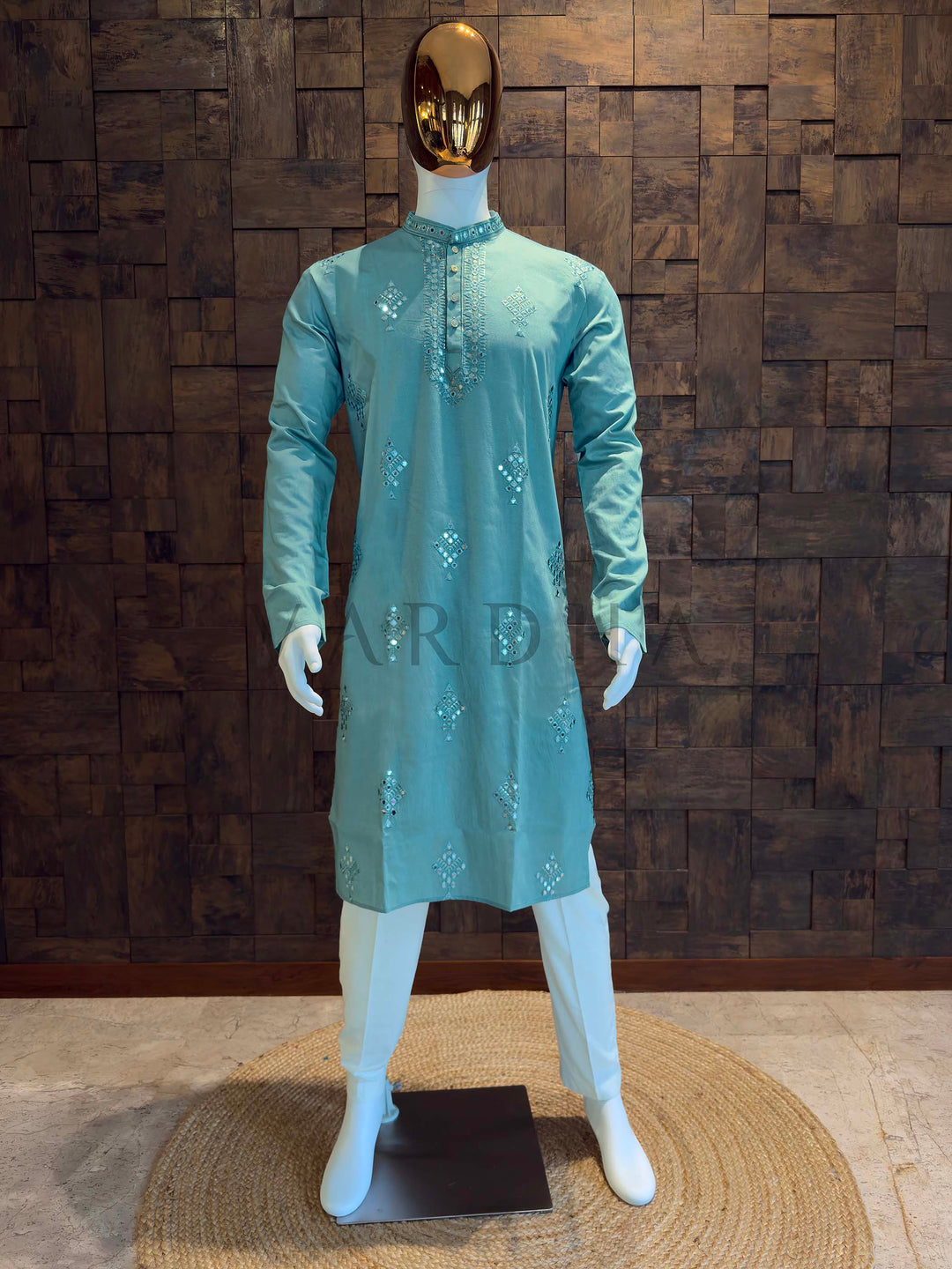 Mannequin wearing a light blue kurta with sequins against a wooden paneled wall. Aqua teal viscose men’s kurta with mandarin collar and mirror-work diamond butis, styled on mannequin with white bottoms for festive and wedding events.