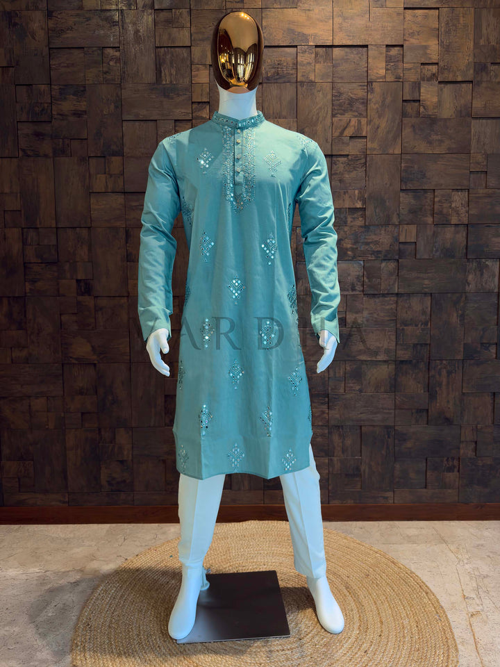 Mannequin wearing a light blue kurta with sequins against a wooden paneled wall. Aqua teal viscose men’s kurta with mandarin collar and mirror-work diamond butis, styled on mannequin with white bottoms for festive and wedding events.