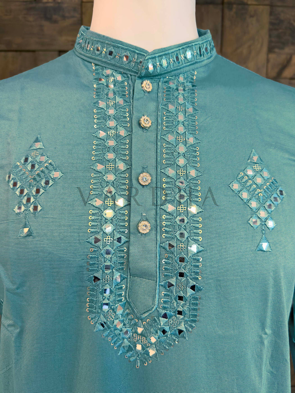 Teal shirt with intricate silver embroidery on a wooden background Close-up of aqua kurta neckline showing mirror-edged band collar and button placket bordered with reflective sheesha embroidery.