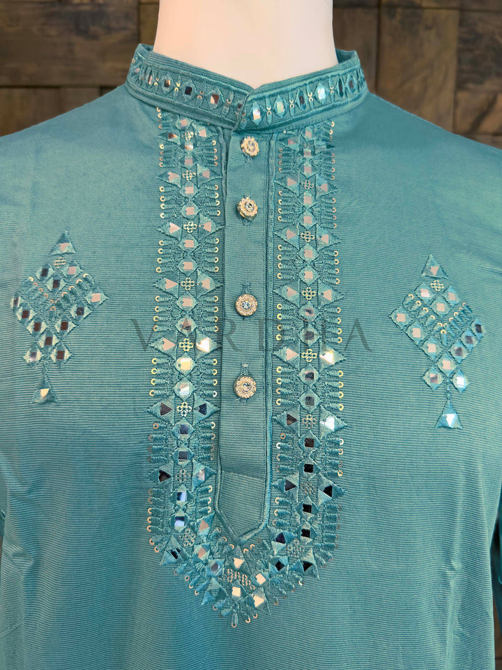 Teal shirt with intricate silver embroidery on a wooden background Close-up of aqua kurta neckline showing mirror-edged band collar and button placket bordered with reflective sheesha embroidery.