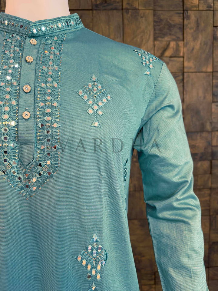 Teal embroidered shirt with intricate patterns on a wooden background Full-length view of teal mirror-work kurta with long sleeves and curved hem, ideal ethnic wear for sangeet and receptions.
