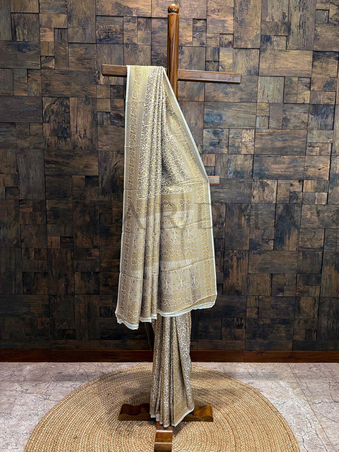 Fabric draped over a wooden stand against a wooden paneled wall. Beige Gold Mysore Silk Saree featuring intricate floral zari weave and traditional border displayed on wooden frame.