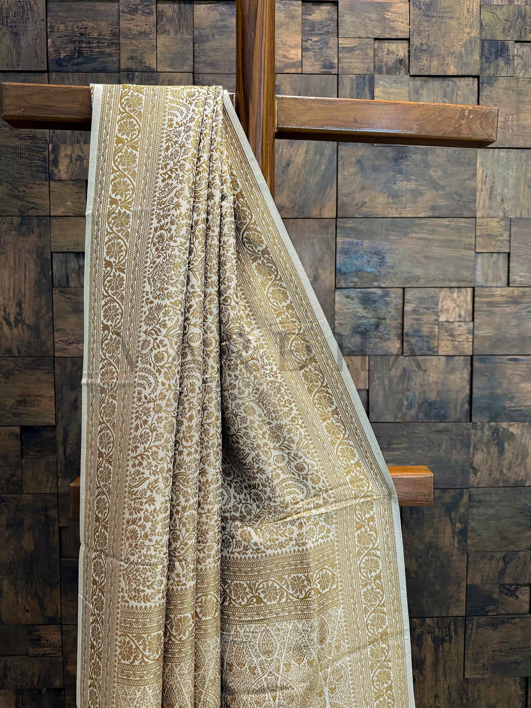 Decorative fabric with intricate patterns draped over a wooden cross against a wooden paneled wall. Pleats of beige-gold viscose Mysore saree showcasing uniform floral motifs and soft texture.
