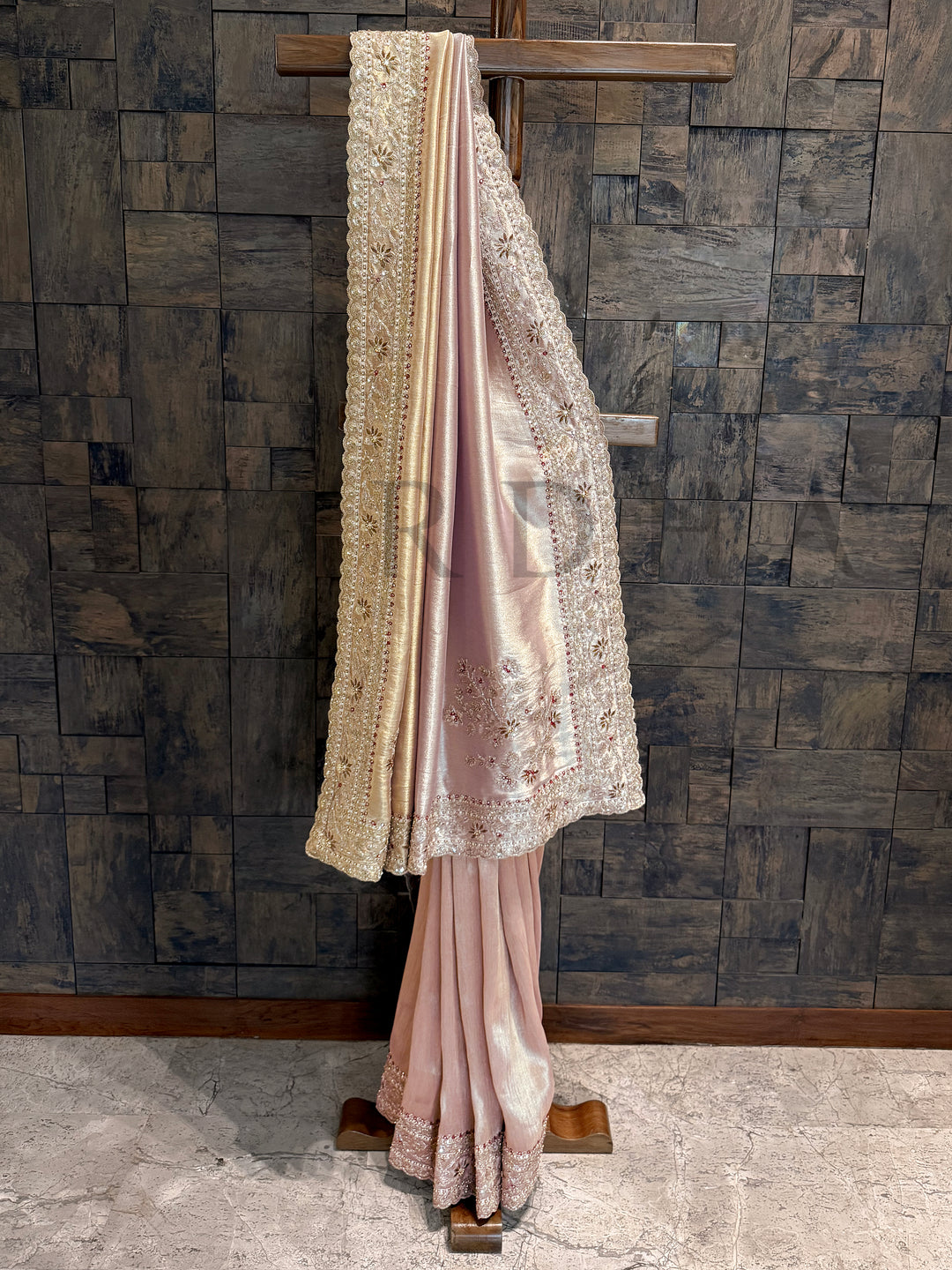 Baby Pink saree with lace details hanging against a textured wall. Cream gold tissue silk saree featuring scalloped borders with intricate hand zardozi embroidery, sequin, and stone detailing.