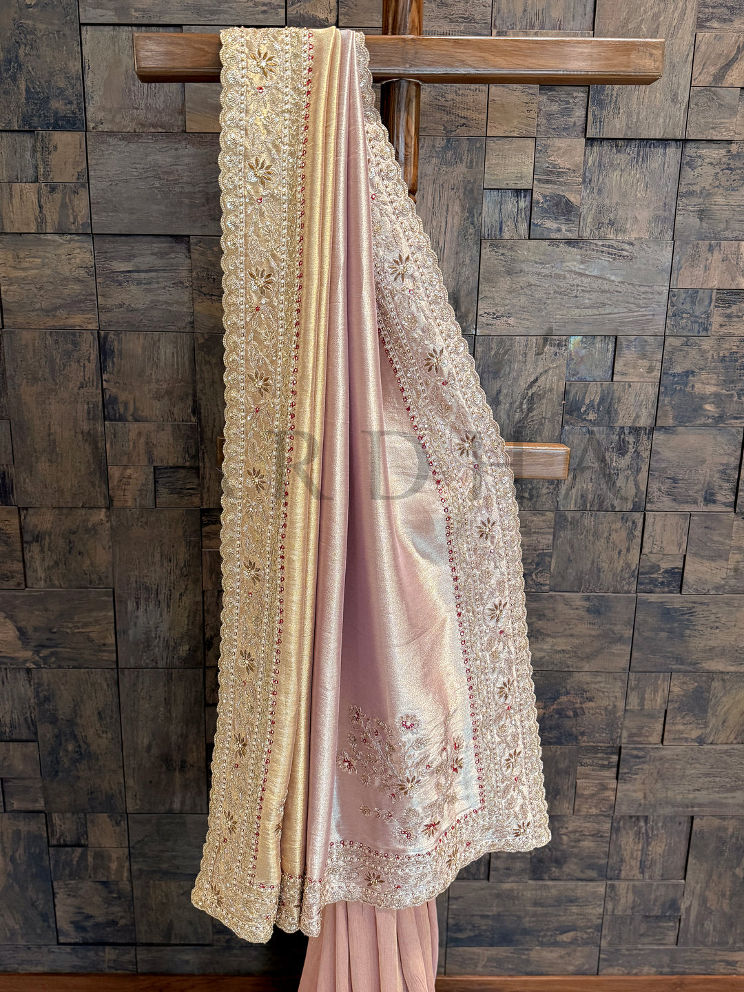 Pink embroidered fabric draped over a wooden rod against a stone wall. Close-up of hand-embroidered border with floral zardozi motifs and red stones on cream tissue silk.