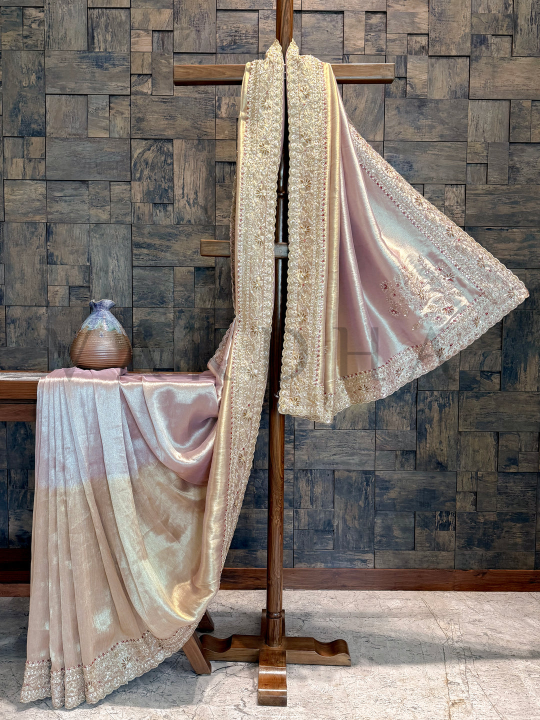 Beige lace fabric draped over a wooden stand against a textured stone wall. Artistic view of saree spread with dual-tone body and intricate embellished edges.