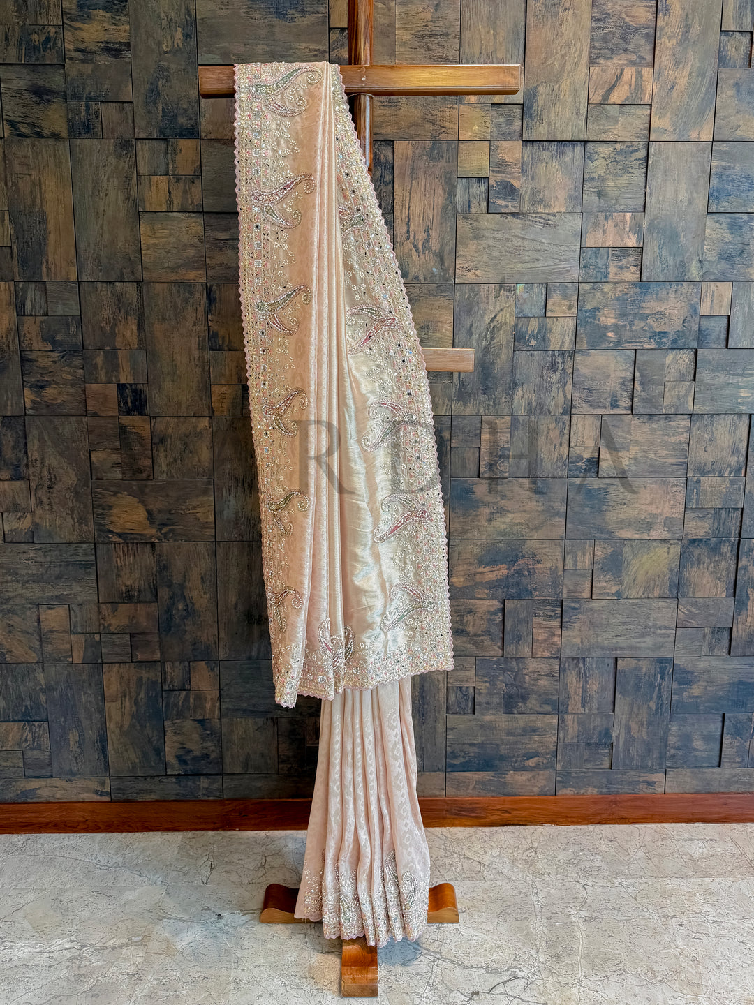 Traditional saree draped over a wooden stand against a textured wall. Baby pink tissue silk saree with hand zardozi embroidery and mirror work border displayed on hanger