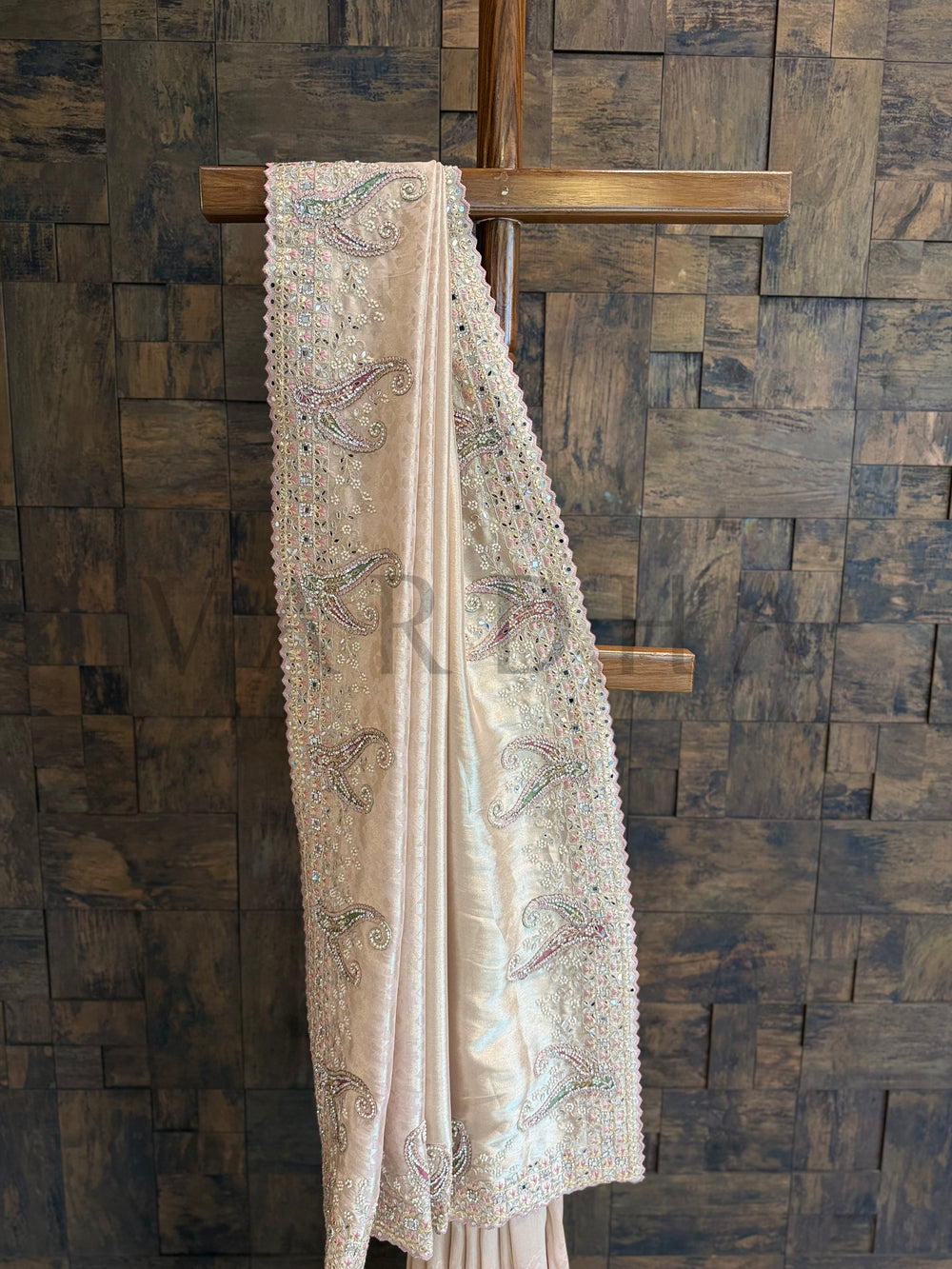Beige embroidered fabric draped over a wooden cross against a wooden paneled wall. Close-up view of pastel paisley motifs hand-embroidered on the soft silk saree