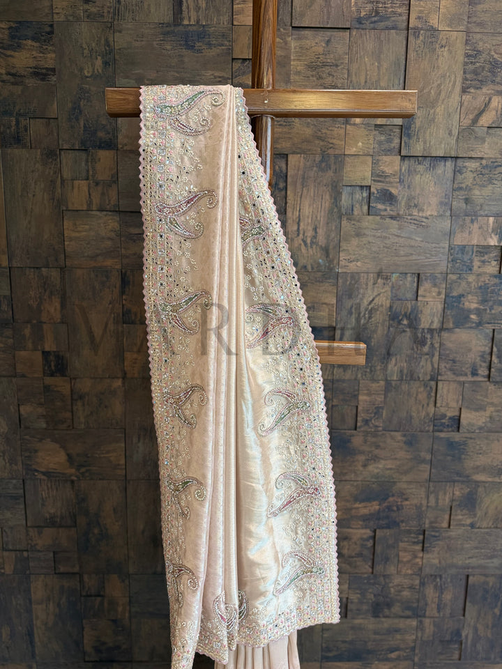 Beige embroidered fabric draped over a wooden cross against a wooden paneled wall. Close-up view of pastel paisley motifs hand-embroidered on the soft silk saree