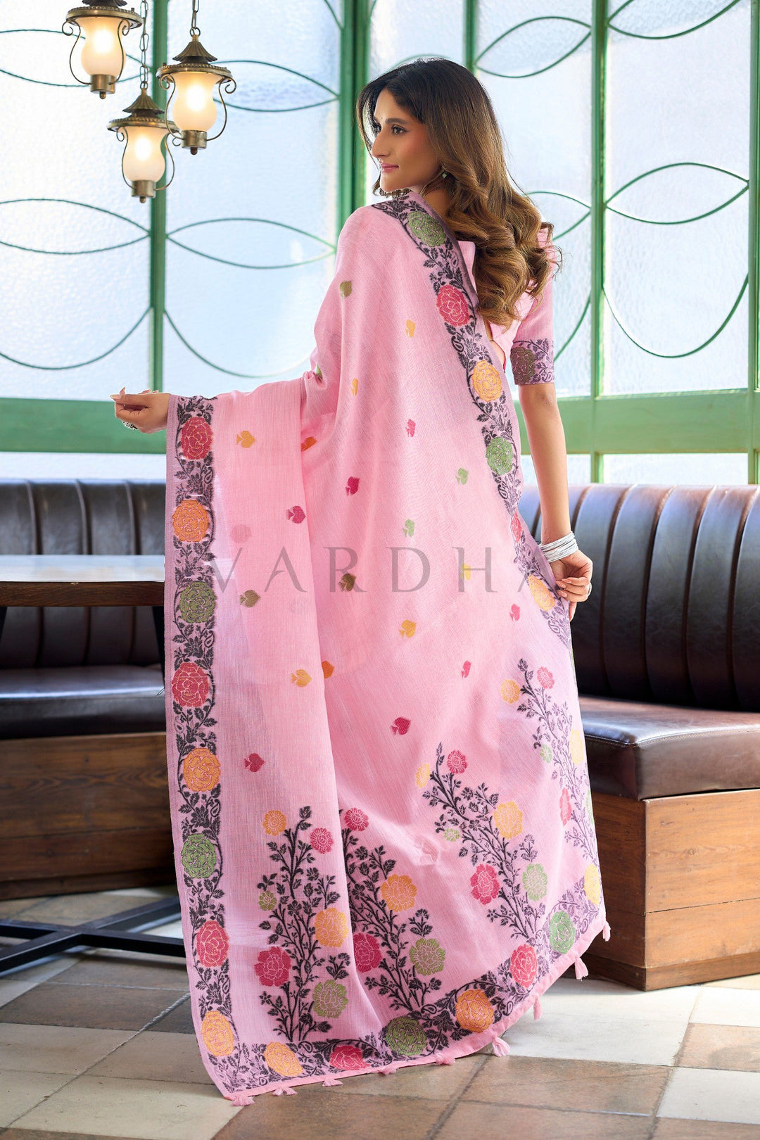 A woman models a light pink saree with colorful floral and paisley border prints in an indoor setting with a bench and green-framed windows in the background. Back view of the baby pink linen jacquard saree, displaying the oversized woven floral vines and matching pink fancy tassels on the designer pallu.