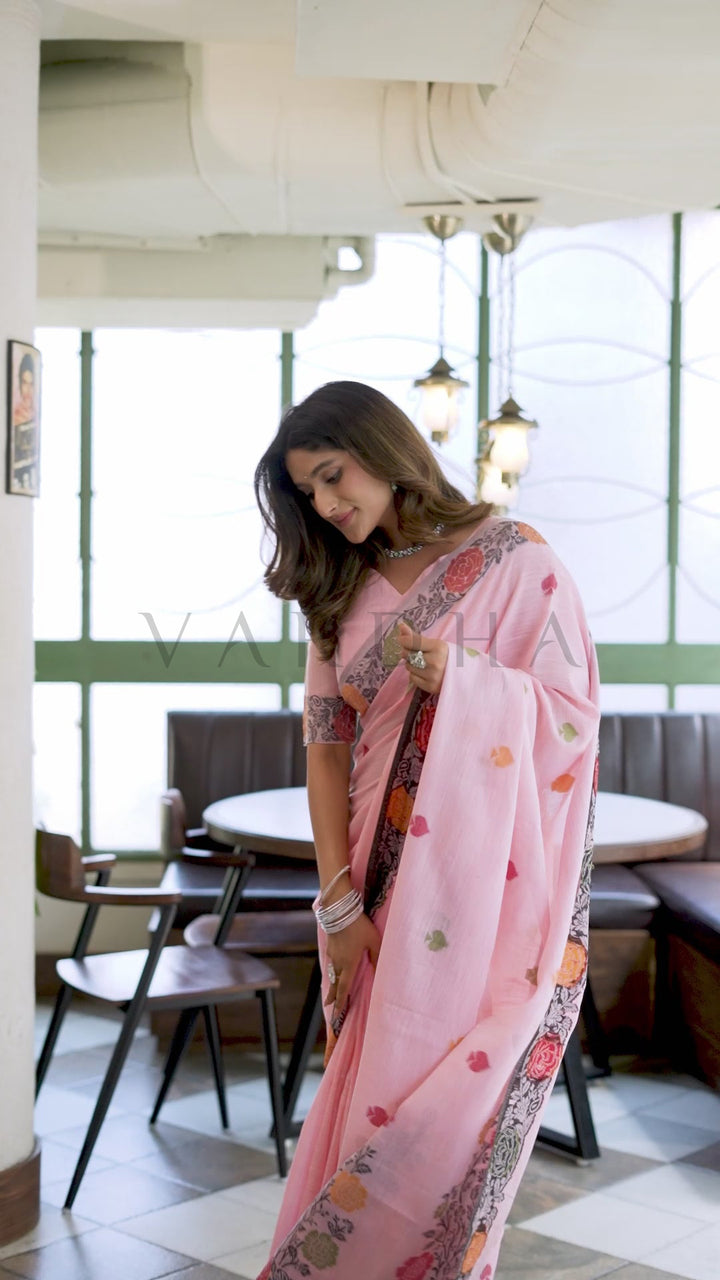 A woman wearing a light pink saree with colorful floral patterns and a matching embroidered blouse, standing in a bright, modern indoor cafe or restaurant setting with tables and chairs in the background. Candid pose of a woman looking down elegantly, showcasing the perfect fall and stunning woven detailing of the baby pink linen jacquard saree.