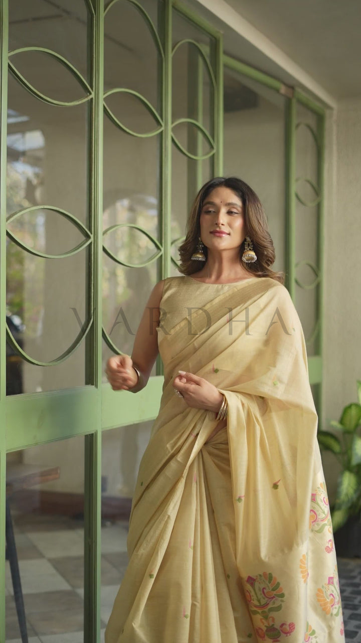 A woman wearing a light beige/cream saree with small gold motifs and floral embroidery near the border, standing in front of a green metal gate. Graceful woman looking down while holding the exquisite Paithani pallu of the champagne beige cotton jacquard saree, showcasing the intricate multicolored threadwork.