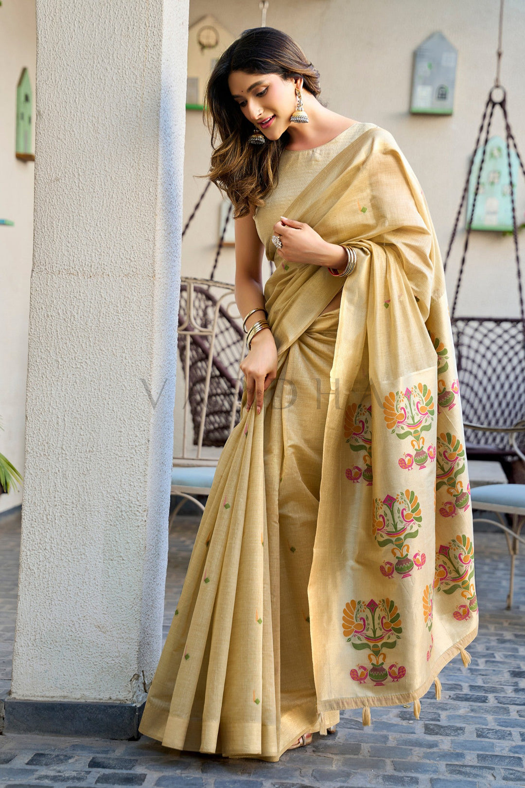 A woman wearing a beige silk saree with colorful floral motifs on the pallu and small gold zari embellishments, standing in a rustic outdoor setting. Beautiful woman posing in the beige cotton jacquard saree, emphasizing the comfortable drape and traditional woven aesthetic perfect for festive occasions.