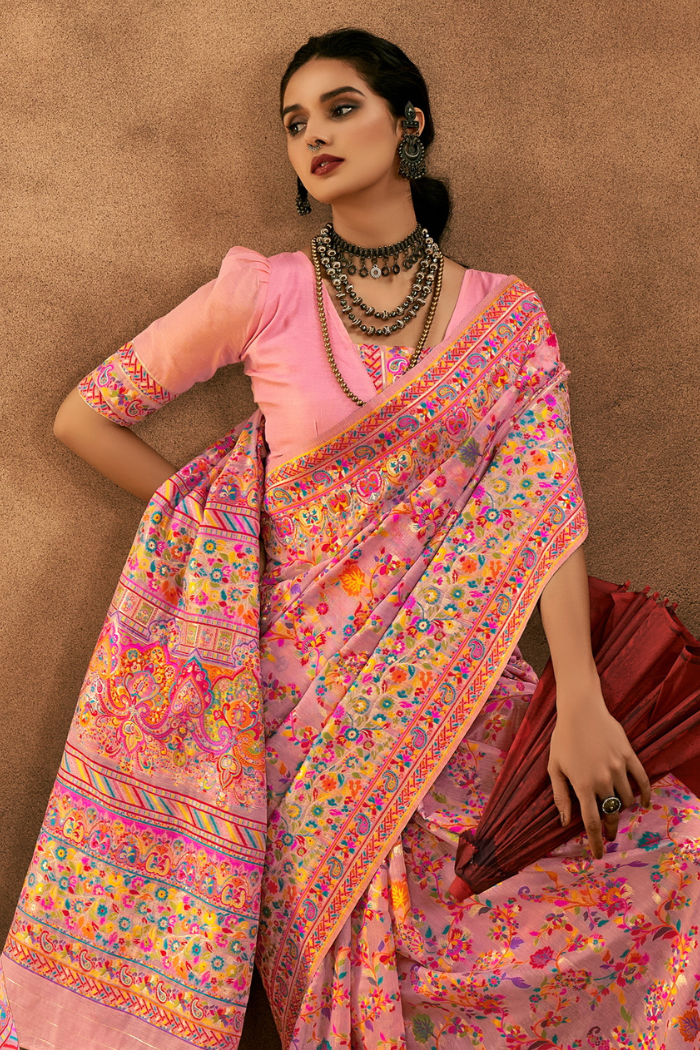 A woman models a pink saree with a heavily embroidered multicolor border and a light pink blouse, posed against a textured brown backdrop. Artistic studio shot focusing on the ornate pashmina jacquard border and traditional jewelry paired with the beige modal cotton saree for wedding wear.