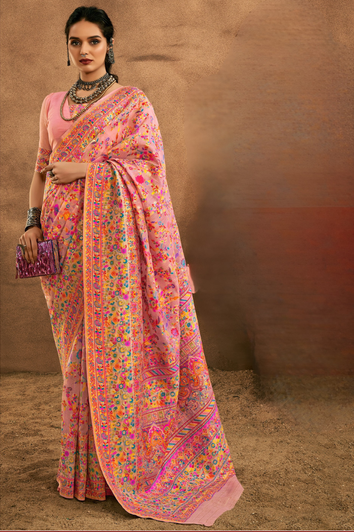 A woman models a pink saree with dense multicolor floral and geometric patterns, paired with a solid pink blouse, against a textured brown studio backdrop. Full-length view of a woman elegantly wearing a beige modal cotton saree with multi-color floral pashmina-style weaving for a luxury festive look.