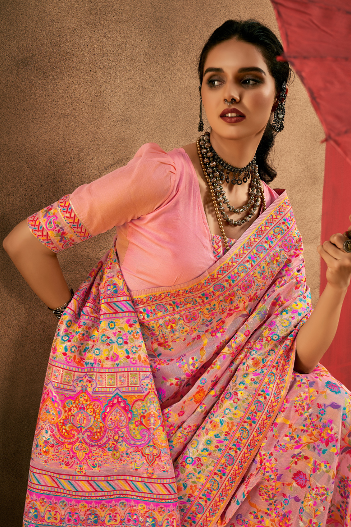 A woman models a pink saree with a heavily embroidered multicolor border and a light pink blouse, posed against a textured brown backdrop. Detailed preview of the beige blouse sleeve featuring ornate multi-colored woven borders in pashmina style.