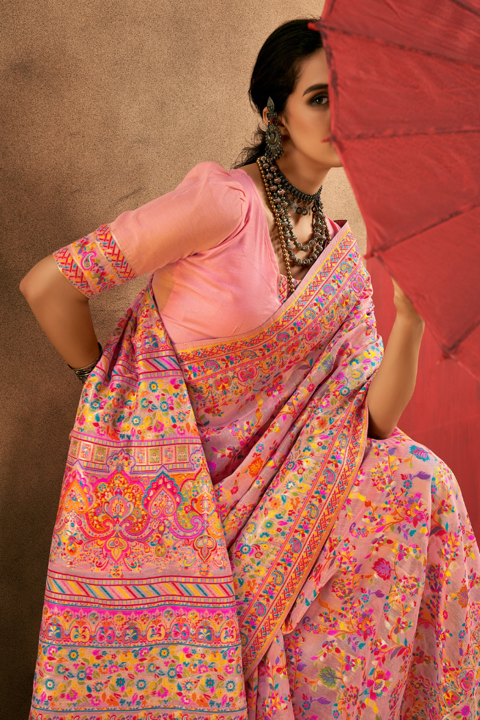 A woman models a pink saree with intricate multicolor floral and geometric patterns, paired with a light pink blouse, holding a red parasol against a textured brown backdrop. Detailed studio shot showing the vibrant pashmina-style floral patterns and the elegant, soft drape of the beige modal cotton saree.