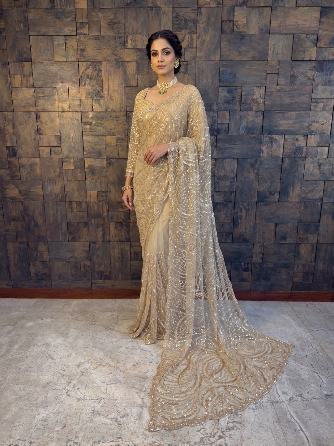 Woman in a sparkling gold saree standing against a textured wall. Beige Net Handwork Saree with Heavy Silver Cutdana Sequin Embroidery.