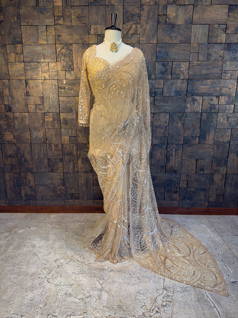 Elegant evening gown on a mannequin against a textured wall. Elegant beige evening gown on a mannequin against a textured wall. House of Vardha Beige Net Saree featuring heavy sparkling embellishments and intricate embroidery, gracefully draped on a mannequin. We offer Ready to wear, Pre drape service, Saree Add on, Custom Stitched Blouse, Fall and Edging, Gift Packaging at Complimentary Price!
