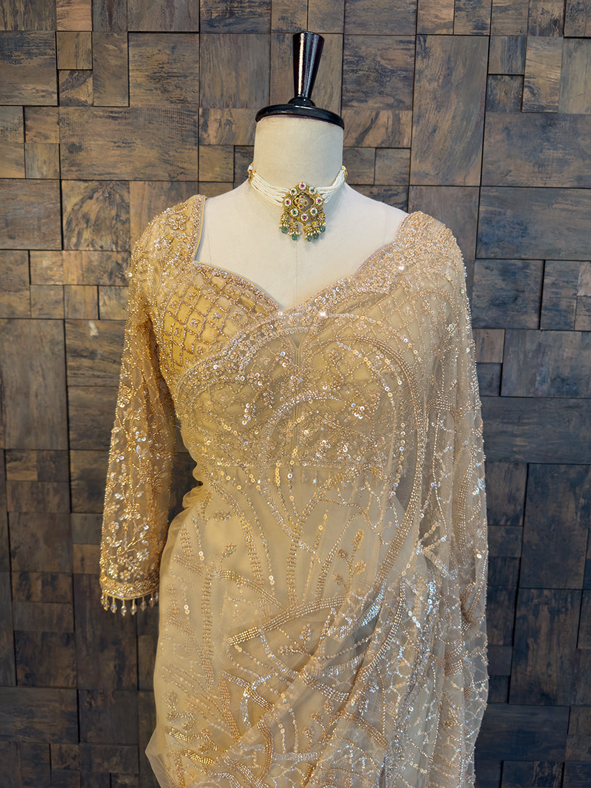 Beige beaded dress on a mannequin against a wooden paneled wall. Beige saree with intricate patterns on a mannequin against a wooden paneled wall. Elegant Beige Net Saree by House of Vardha showcasing exquisite all-over sequin work and a matching embellished blouse. We offer Ready to wear, Pre drape service, Saree Add on, Custom Stitched Blouse, Fall and Edging, Gift Packaging at Complimentary Price!
