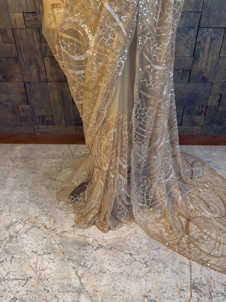 Gold and silver patterned fabric draped over a wooden wall. Close-up view of the House of Vardha Beige Net Saree highlighting the detailed sparkling embroidery and delicate sheer fabric. We offer Ready to wear, Pre drape service, Saree Add on, Custom Stitched Blouse, Fall and Edging, Gift Packaging at Complimentary Price!