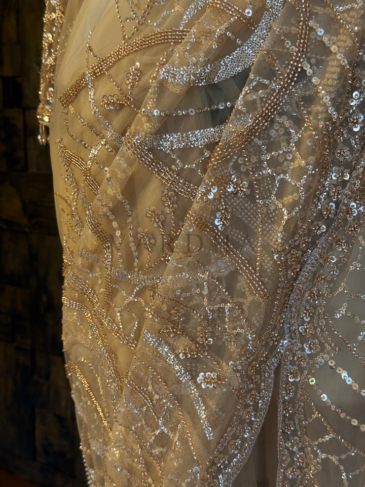 Close-up of a gold and silver beaded fabric with intricate patterns. Beautifully draped Beige Net Saree highlighting the elegant sheer fall of the fabric and shimmering silver-toned embroidery by House of Vardha. We offer Ready to wear, Pre drape service, Saree Add on, Custom Stitched Blouse, Fall and Edging, Gift Packaging at Complimentary Price!