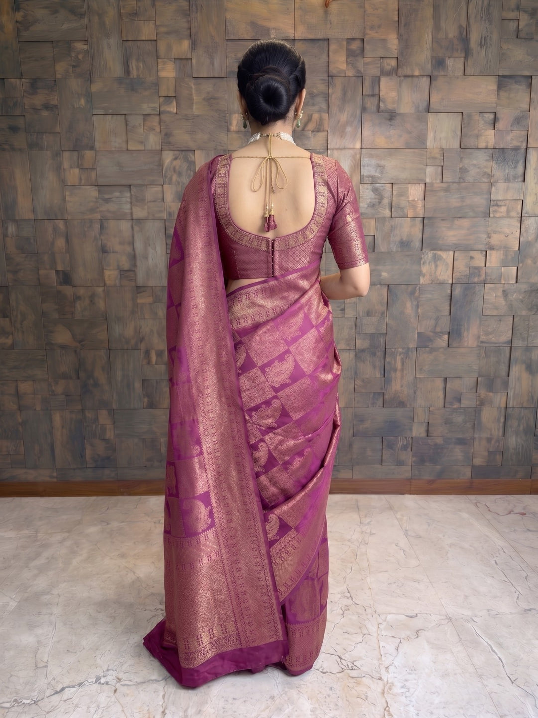 A woman viewed from behind wearing a purple and gold brocade saree with a matching blouse, standing against a rustic wooden paneled wall. A full-length side-angle shot of a woman stepping forward while wearing a berry purple Kanjivaram silk saree. The pose captures the elegant drape, movement of the fabric, and the prominent gold zari borders.