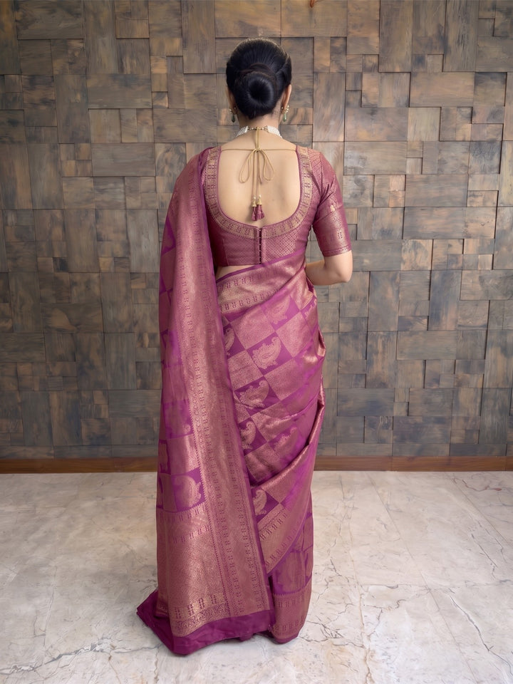 A woman viewed from behind wearing a purple and gold brocade saree with a matching blouse, standing against a rustic wooden paneled wall. A full-length side-angle shot of a woman stepping forward while wearing a berry purple Kanjivaram silk saree. The pose captures the elegant drape, movement of the fabric, and the prominent gold zari borders.