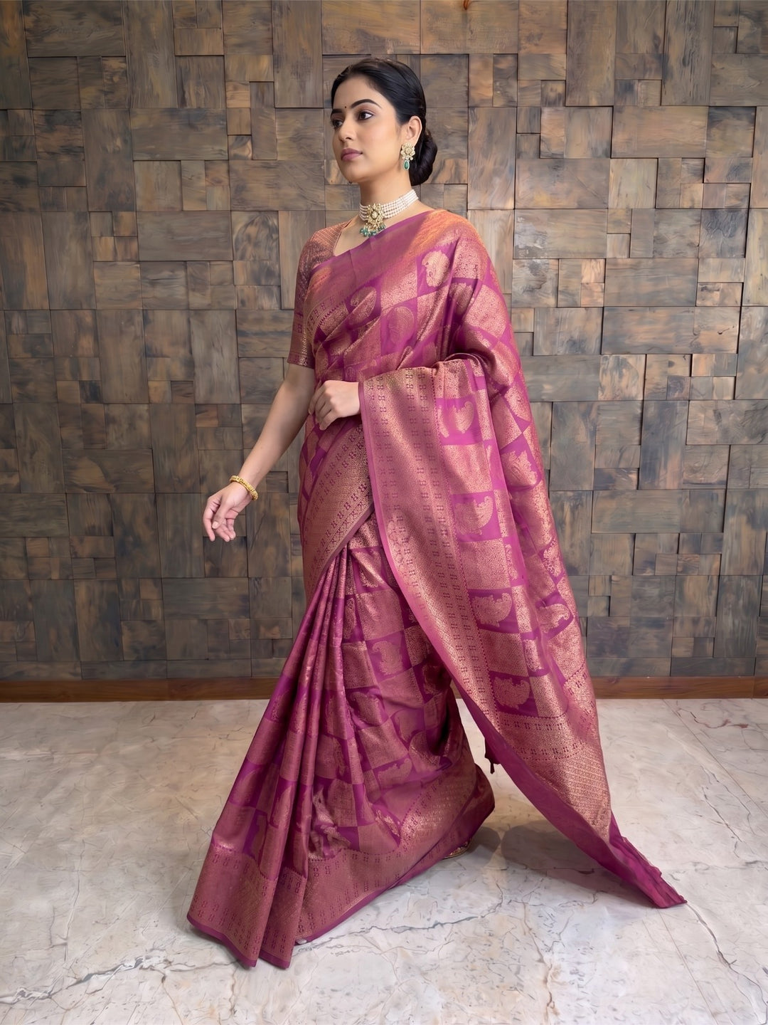 A woman models a magenta pink and gold Zari woven saree with geometric patterns, standing against a rustic wooden paneled wall. A full-length side-angle shot of a woman stepping forward while wearing a berry purple Kanjivaram silk saree. The pose captures the elegant drape, movement of the fabric, and the prominent gold zari borders.