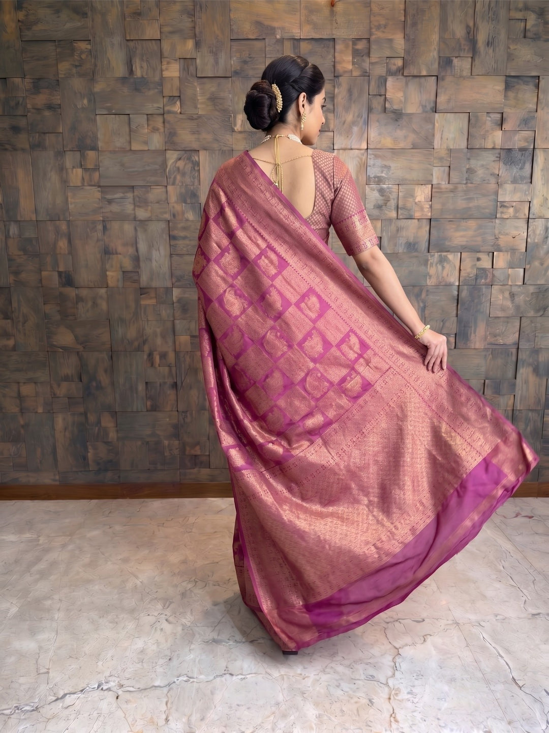 A woman wearing a dusty rose pink saree with a magenta geometric pattern and a matching blouse, standing in front of a rustic wooden wall. A full-length back view of a woman in a berry purple Kanjivaram silk saree, looking slightly over her shoulder. She is holding the edge of the saree&