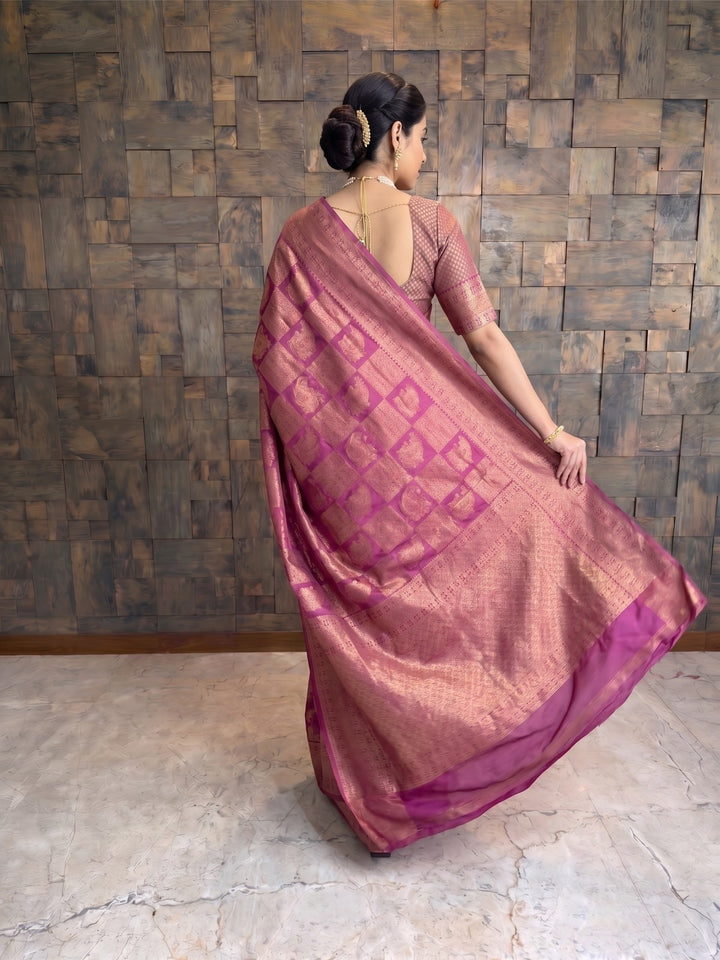 A woman wearing a dusty rose pink saree with a magenta geometric pattern and a matching blouse, standing in front of a rustic wooden wall. A full-length back view of a woman in a berry purple Kanjivaram silk saree, looking slightly over her shoulder. She is holding the edge of the saree&
