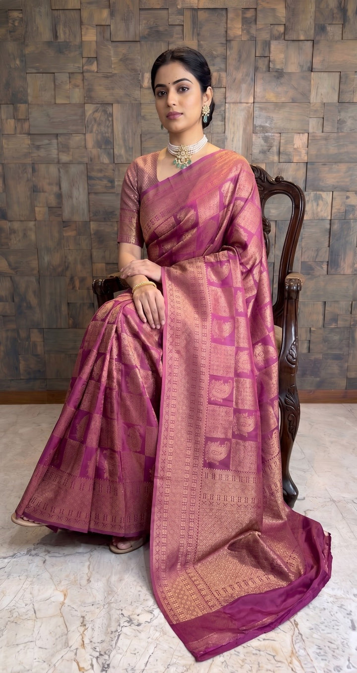A woman seated on a dark wooden chair wearing a pink and gold zari-embellished silk saree, posed against a textured stone wall. A full-length shot of a woman seated elegantly on a carved wooden chair. She is wearing a berry purple Kanjivaram silk saree with gold zari, draped beautifully around her with the pallu cascading toward the floor.