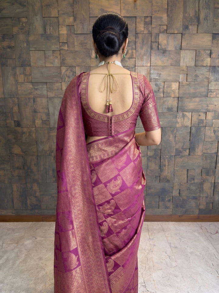 A woman viewed from behind wearing a magenta purple silk saree with gold zari work and a matching embroidered blouse, standing against a textured brown wooden wall. A full-length shot of a woman seated elegantly on a carved wooden chair. She is wearing a berry purple Kanjivaram silk saree with gold zari, draped beautifully around her with the pallu cascading toward the floor.