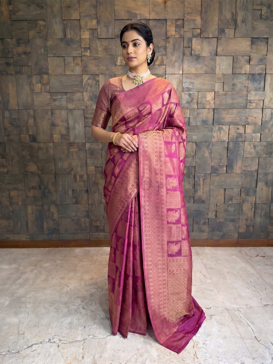 A full-length front view of a woman standing and posing in a berry purple Kanjivaram silk saree featuring gold zari checkered and paisley patterns. She has her hands softly clasped in front of her and is wearing a matching short-sleeved blouse, pearl and emerald jewelry, and her hair is styled in a sleek low bun. A woman modeling a magenta pink and gold Banarasi silk saree with geometric patterns, standing against a rustic wooden paneled wall.