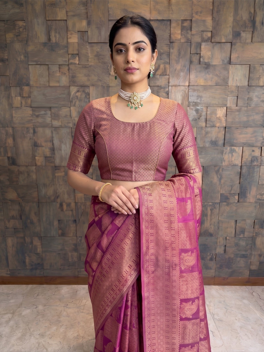 A woman modeling a traditional Indian saree with a matching blouse. The saree is a deep magenta-purple with gold zari woven patterns, paired with a matching short-sleeve blouse in a dusty rose pink with gold embroidery. A three-quarter length front view of a woman wearing a berry purple Kanjivaram silk saree. The closer angle highlights the detailed gold zari patterns on the blouse and saree border, as well as her pearl and emerald choker necklace.