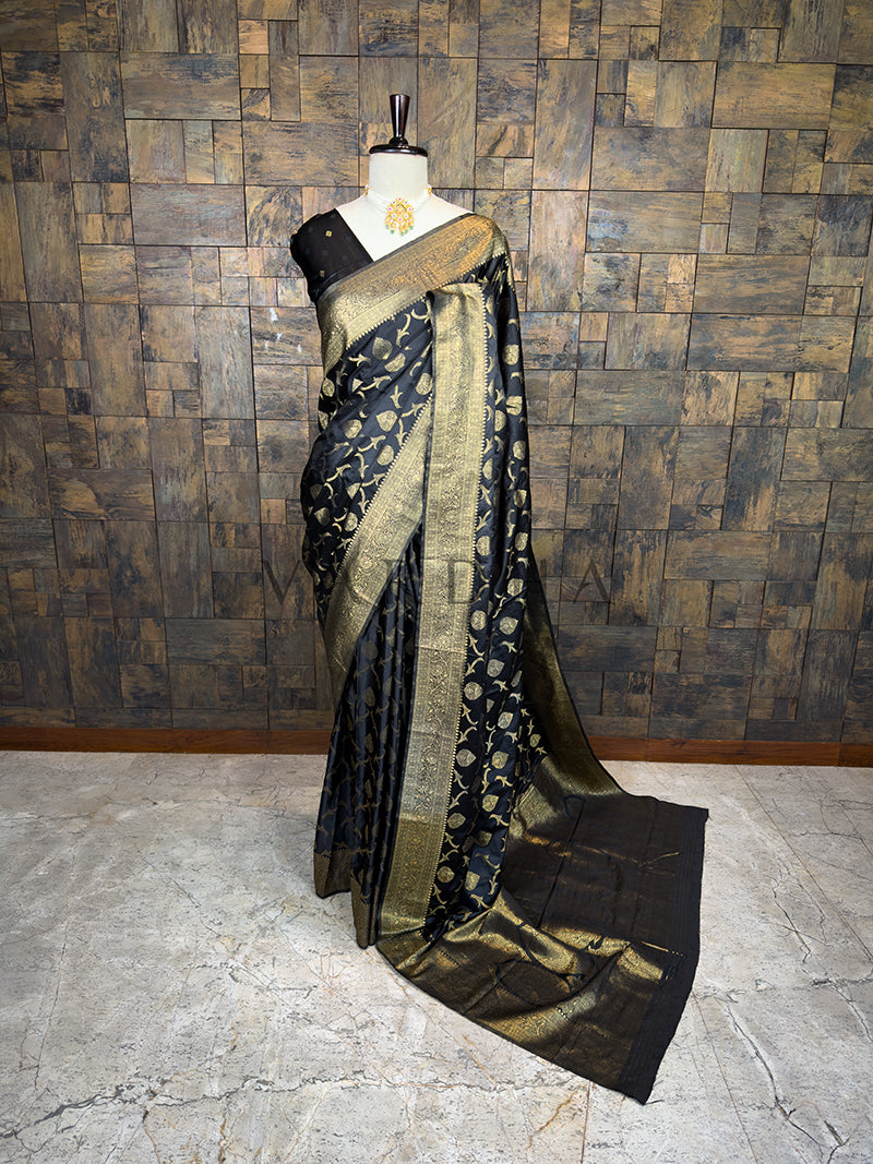 Black and gold saree on a mannequin against a textured wall. House of Vardha Black Banarasi Satin Silk Saree featuring Gold Zari leaf motifs and a matching border, elegantly draped on a mannequin. We offer Ready to wear, Pre drape service, Saree Add on, Custom Stitched Blouse, Fall and Edging, Gift Packaging at Complimentary Price!