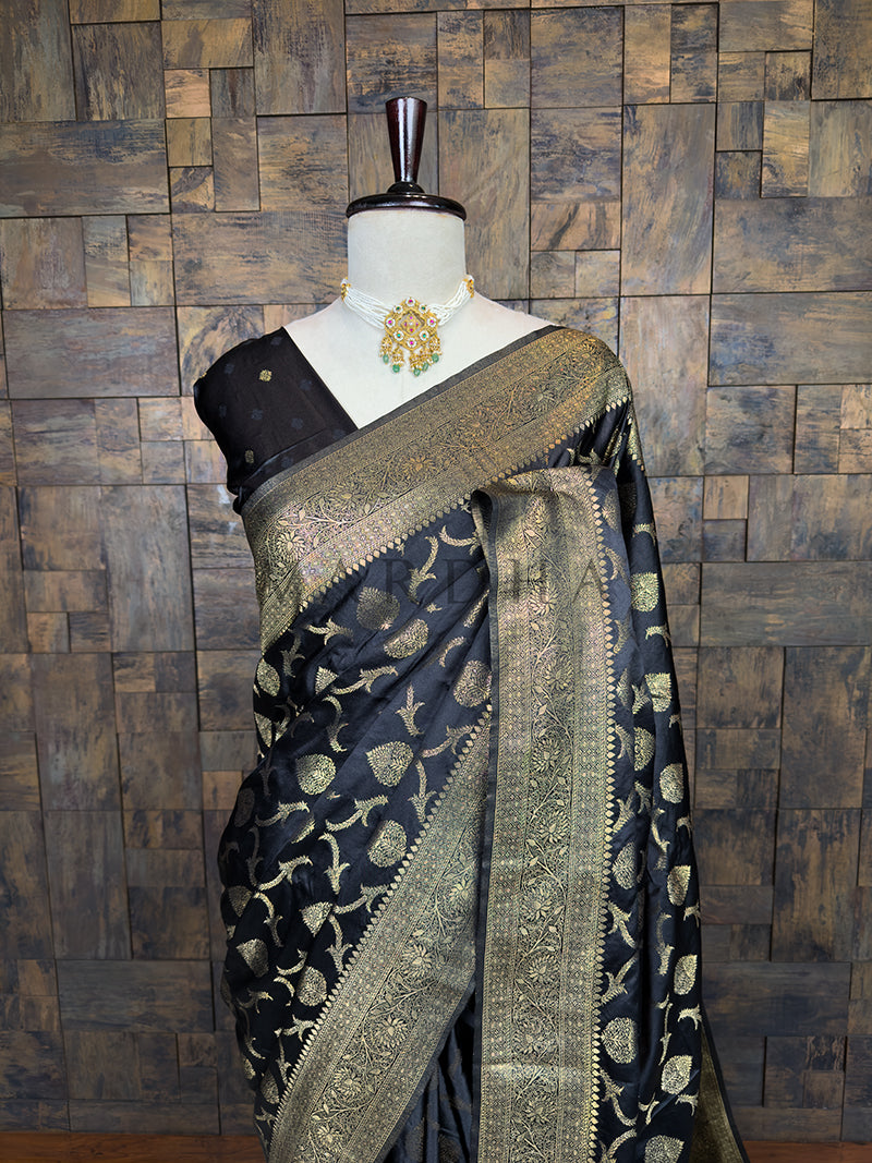 Black saree with gold patterns on a mannequin against a wooden paneled wall. Lustrous Black Banarasi Satin Silk Saree by House of Vardha showcasing intricate Gold Zari weaving and a rich, smooth texture. We offer Ready to wear, Pre drape service, Saree Add on, Custom Stitched Blouse, Fall and Edging, Gift Packaging at Complimentary Price!