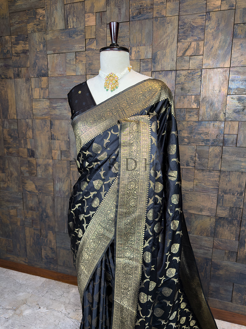 Black saree with gold patterns on a mannequin against a wooden paneled wall. Close-up view of the House of Vardha Black Banarasi Satin Silk Saree highlighting the detailed Gold Zari motifs and the beautifully woven traditional border. We offer Ready to wear, Pre drape service, Saree Add on, Custom Stitched Blouse, Fall and Edging, Gift Packaging at Complimentary Price!