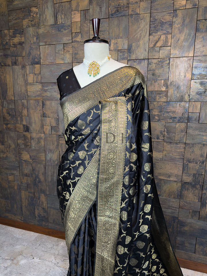 Black saree with gold patterns on a mannequin against a wooden paneled wall. Close-up view of the House of Vardha Black Banarasi Satin Silk Saree highlighting the detailed Gold Zari motifs and the beautifully woven traditional border. We offer Ready to wear, Pre drape service, Saree Add on, Custom Stitched Blouse, Fall and Edging, Gift Packaging at Complimentary Price!